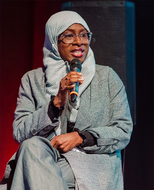 An image of Black person speaking into a microphone looking at someone to their left, off camera.