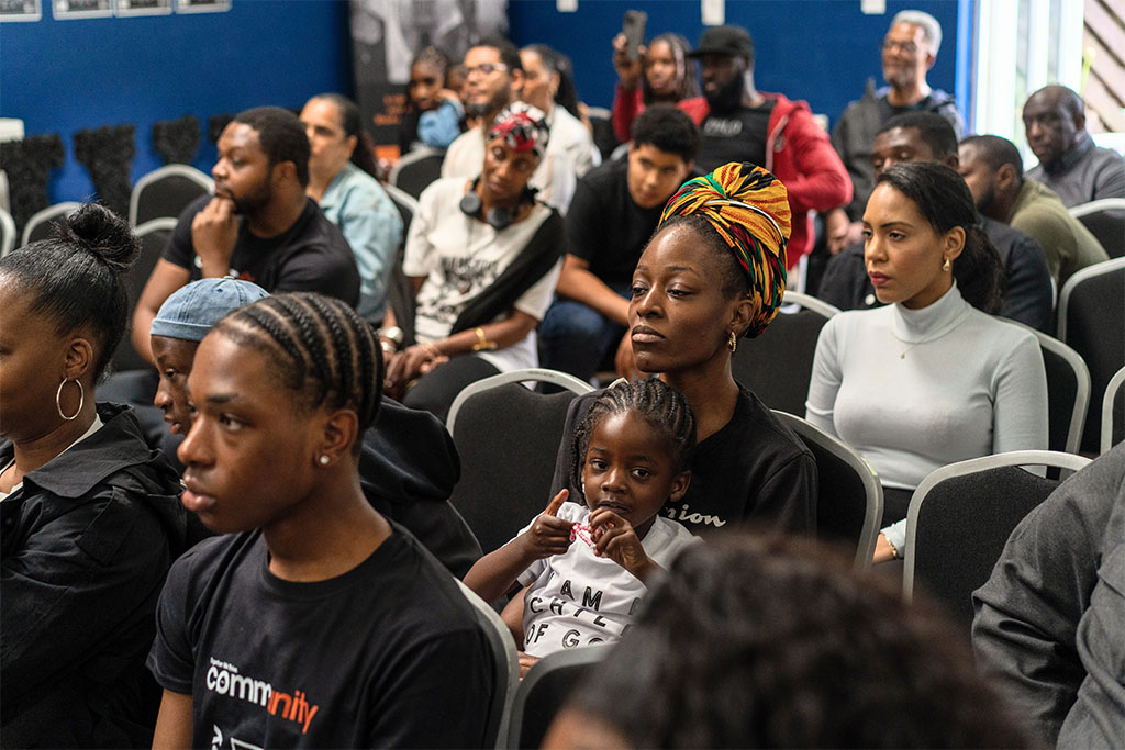 An image of a group of predominantly Black people sat in a room, watching something in unison.