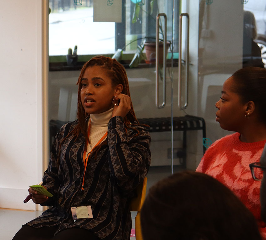 An image of two Black people sat down, the person on the right is speaking to someone in front of them, out of view of the camera.