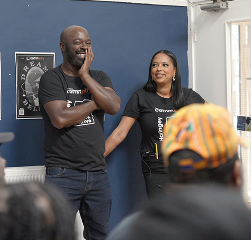 An image of two Black people stood next to a blue wall smiling.