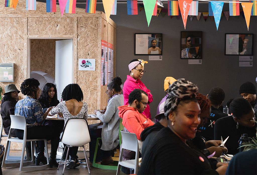 An image of groups of Black people sat at tables engaged in discussions. A Black person wearing a bright pink top, a colourful bandana, and glasses, is stood in the centre of the room.