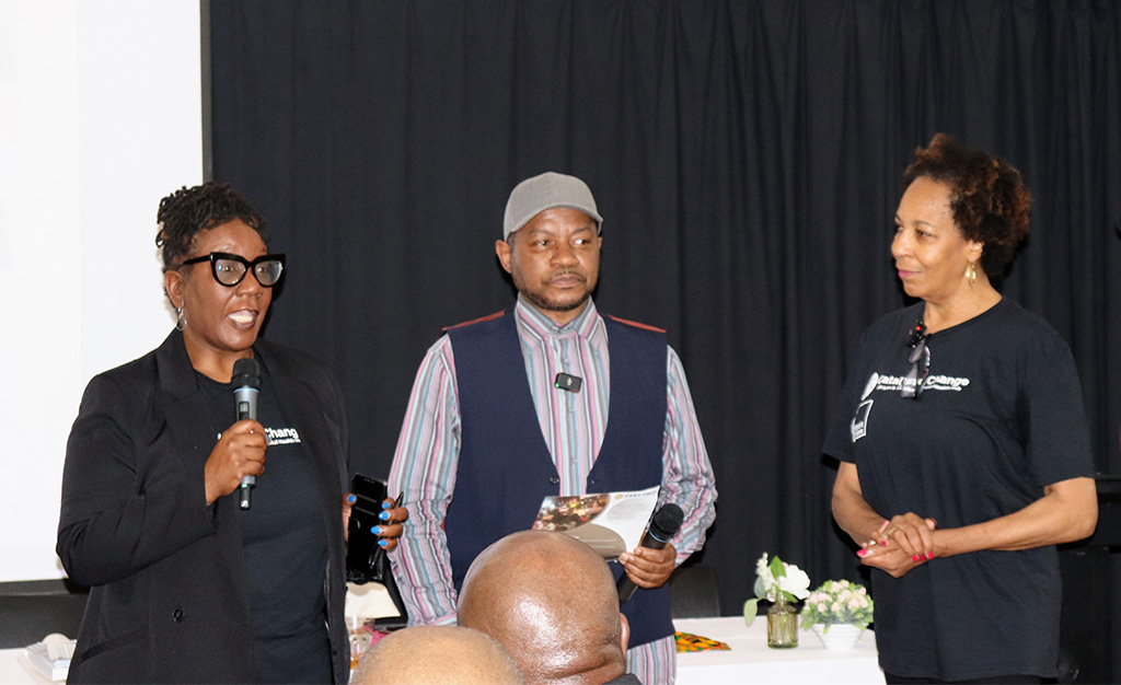 An image of three Black people stood against a black curtain. The person on the far left is talking into a microphone while addressing the audience.